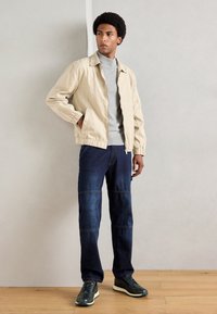 Homem vestido com um casaco bege, suéter de gola alta cinza, jeans de retalhos escuros e tênis pretos, em pé sobre um piso de madeira contra uma parede cinza.