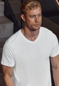 Bílá bavlněná polo tričko s výstřihem ve tvaru V, krátkými rukávy, hladkou texturou, přiléhavým střihovým designem, nošené mužem s krátkými blond vlasy v moderním vnitřním prostředí.