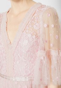Robe rose brodée avec un décolleté en V plongeant, des manches en dentelle florale transparente, des accents en tulle et une ceinture en satin. Motifs floraux délicats sur toute la surface.
