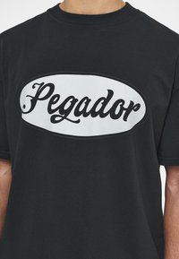 Černé bavlněné tričko s velkým bílým oválným motivem a textem "Pegador" v tučném černém písmu přes hrudník. Krátké rukávy.