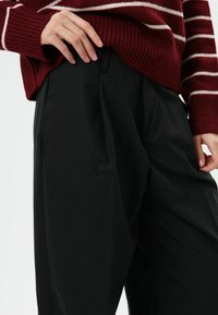 Zwarte broek met een losse pasvorm, met een gladde textuur en wijde broekspijpen. Een bordeauxrood gestreepte trui bedekt gedeeltelijk de tailleband.