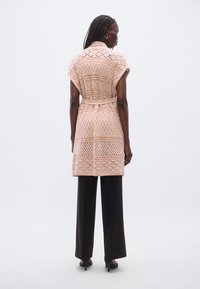 Πλεκτή ζακέτα σε ανοιχτό ροδακινί με υφασμένα μοτίβα, κοντά μανίκια και δεμένη ζώνη στη μέση, φορεμένη πάνω από μαύρο παντελόνι με φαρδιά πόδια.