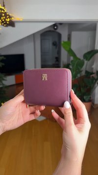 Πορτοφόλι από δέρμα μπορντό με μεταλλικά στοιχεία σε χρυσό τόνο, με λογότυπο στο μπροστινό μέρος, κλείσιμο με φερμουάρ και λεία υφή. Συμπαγής και ορθογώνιο σχήμα.