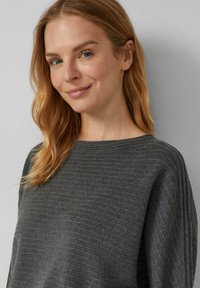 Femme blonde aux yeux bleus souriant légèrement, portant un pull côtelé gris foncé, se tenant contre un mur gris clair.