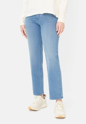 Person iført lyseblå jeans med lige ben og hvide sneakers, stående mod en ensfarvet lys baggrund.