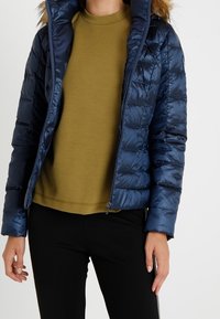 Laivastonsininen toppatakki, jossa on pystysuora tikkauskuvio, turkiskaistaleinen huppu, vetoketjukiinnitys ja joka on puettu oliivinvihreän paidan päälle.