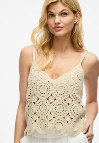 Top de ganchillo en beige con un dobladillo en picos, que presenta patrones circulares y tirantes delgados ajustables. Textura ligera y de punto abierto.