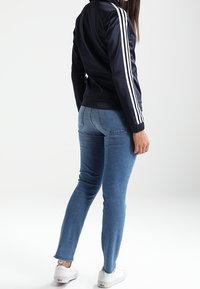 Chaqueta deportiva azul marino con rayas blancas, diseño ajustado. Jeans skinny azul claro, dobladillo ligeramente deshilachado, combinados con zapatillas blancas.