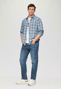 Lichtblauw geruit overhemd over een witte graphic t-shirt, gecombineerd met blauwe spijkerbroeken en witte sportschoenen met donkere accenten.