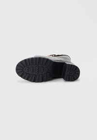 Semelle de botte noire avec un motif de rainure profond, présentant une forme arrondie, en matériau caoutchouc, et des accents en métal doré sur le côté.