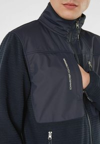Giacca softshell navy con colletto alto zip, caratterizzata da un motivo a maglia testurizzato e pannelli lisci, include zip e dettagli del logo.