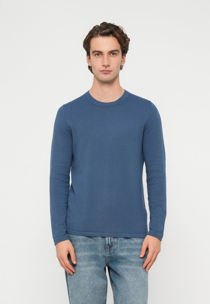 Niebieska koszulka z długim rękawem, o okrągłym dekolcie, z prążkowanym materiałem, dopasowanym krojem i prostym dołem, noszona z jasnoniebieskimi dżinsami.