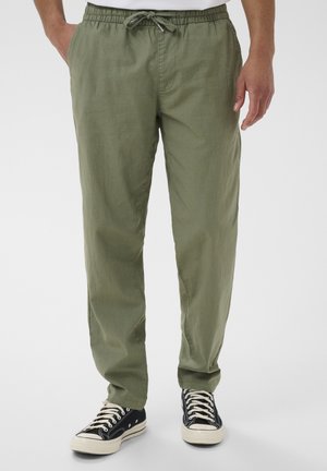 Mand iført olivengrønne snorebukser og sorte lave canvas-sneakers, stående mod en ensfarvet baggrund.