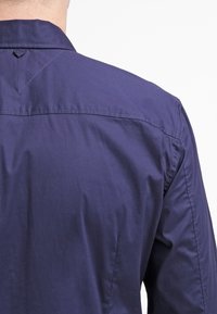 Granatowa koszula z klasycznym kołnierzem, gładkim materiałem i trójkątnym przeszyciem na tylnej części karczka. Dopasowany krój.