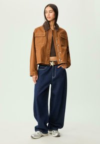 Ung kvinde iført en brun cropped jakke, mørkebrun top, brede mørkeblå jeans med et stjernebælte og sølvfarvede sneakers, stående mod en lys baggrund.