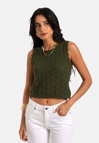 Grüner gestrickter Crop-Top mit Diamantmustern, gewellten Kanten und einer strukturierten Oberfläche, kombiniert mit weißen, hoch taillierten Hosen.