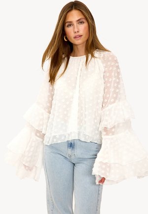 Blouse blanche transparente avec manches évasées à volants, tissu texturé avec motifs floraux, encolure ronde et coupe ample. Associée à un jean en denim.