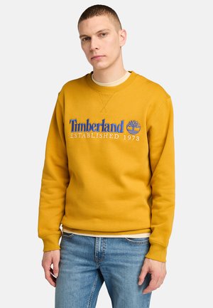 Geel sweatshirt met geborduurde blauwe "Timberland" tekst en logo. Beschikt over een ronde hals, ribgebreide manchetten en een casual pasvorm. G gedragen met blauwe jeans.