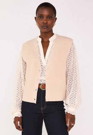 Femme portant un gilet beige en tricot sur un chemisier en dentelle florale blanc à manches bouffantes, jeans foncés, ceinture marron et boucles d'oreilles dorées en créole, debout.