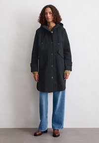 Schwarzer wasserdichter Mantel mit Kapuze, ausgestattet mit zwei Fronttaschen, einem Knopfverschluss und gerippten Bündchen. Kombiniert mit hellblauen weit geschnittenen Jeans.