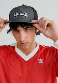 Μαύρο και γκρι καπέλο με το λογότυπο "ADIDAS", κρατημένο από ένα άτομο. Η κόκκινη φανέλα έχει κάθετες ρίγες και λευκές λεπτομέρειες στον γιακά.