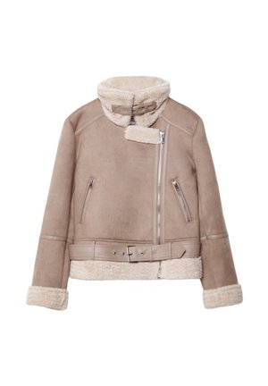 Persoon draagt een beige schapenvachtjas, wit overhemd, blauwe wijdere spijkerbroek en bruine suède schoenen, staand tegen een effen achtergrond.