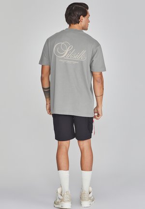 Camiseta de algodón gris con un gran logo crema en la espalda, combinada con pantalones cortos negros. El modelo lleva calcetines blancos y zapatillas beiges.