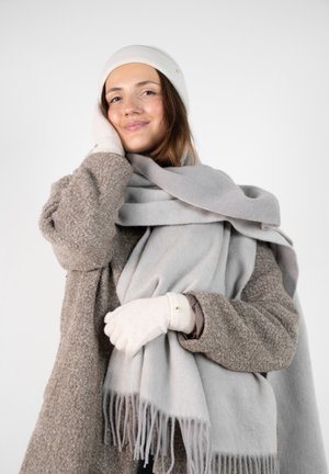 Light gray wool scarf with fringed edges, layered over a textured brown coat. Model wears a cream hat and gloves, with subtle gold accents.
