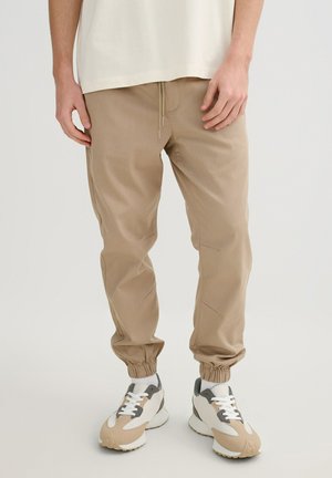 Hombre con pantalones jogger beige con puños elásticos y zapatillas beige, blancas y grises sobre un fondo liso.