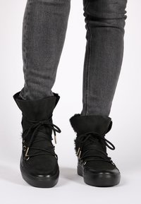 Botines negros hasta el tobillo con ribete de piel, material de ante, punta redonda, diseño con cordones y detalles de herrajes dorados. Usados con jeans ajustados grises.