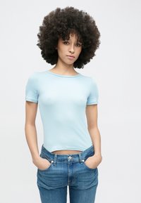 Világoskék, bordázott rövid ujjú felső kerek nyakkivágással, magas derekú kék farmerral párosítva. A modell zsebre tett kézzel pózol.