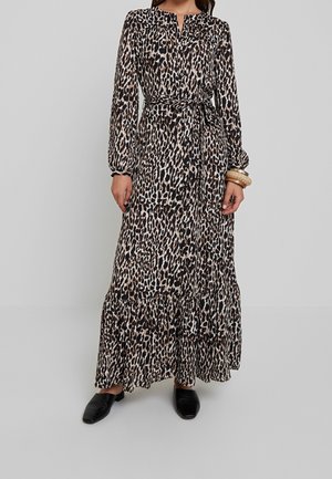 Femme portant une robe longue à manches longues imprimée léopard avec une taille nouée et des chaussures noires à enfiler sur un fond uni.