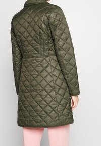 Quiltad lång jacka i olivgrön, med ett diamantmönster, krage och figursydd midja. Slät textur och sömdetaljer synliga.