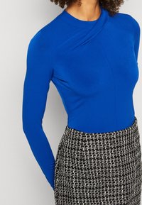 Blå långärmad topp med hög krage och draperad design, tillsammans med en strukturerad svart och vit tweedkjol med geometriskt mönster.