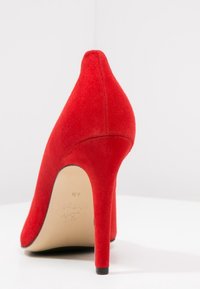 Scarpa con tacco alto in suede rosso, con punta affusolata e tacco a stiletto. Texture liscia; vista posteriore che mette in mostra altezza e forma.