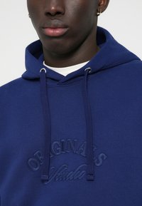 Haut à capuche bleu marine en tissu mélangé de coton, avec logo brodé "ORIGINALS" et cordons de serrage. Coupe oversize avec une texture douce.