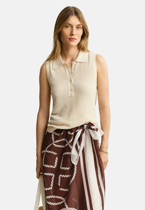 Femme portant un polo tricoté beige sans manches et une jupe portefeuille marron avec des motifs de corde blanche et de rayures, debout devant un fond uni.