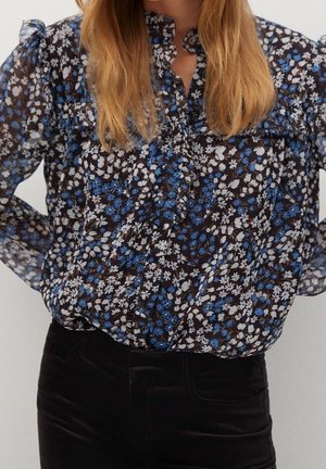 Frau trägt eine durchsichtige schwarze Bluse mit blauem und weißem Blumenmuster, ausgestattet mit Rüschen und gepufften Schultern, kombiniert mit einer schwarzen Hose.