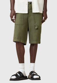 Pantaloni cargo verde oliva con molte tasche, vestibilità rilassata e orlo dritto, abbinati a sandali neri a scivolo e calze bianche a metà polpaccio.