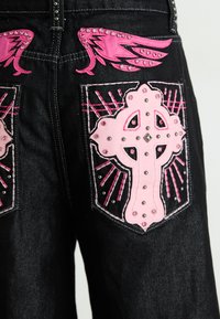 Crne traperice s ružičastim križem na stražnjim džepovima, ukrašene rhinestones i šiljcima; poboljšane ružičastim šavovima.