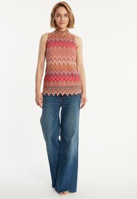 Mehrfarbiges ärmelloses Oberteil mit Zickzackmuster, hohem Kragen und gewelltem Saum. Kombiniert mit weiten blauen Jeans.