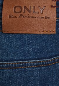 Etiqueta de couro castanho com texto em relevo "ONLY" e "Blue Denim anno 1995" presa a um tecido de denim azul, apresentando costura dourada e textura.