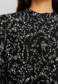 Svart blus med ett vitt blommönster och en instoppad halsringning. Lättviktig tyg med en subtil textur och flödande design.