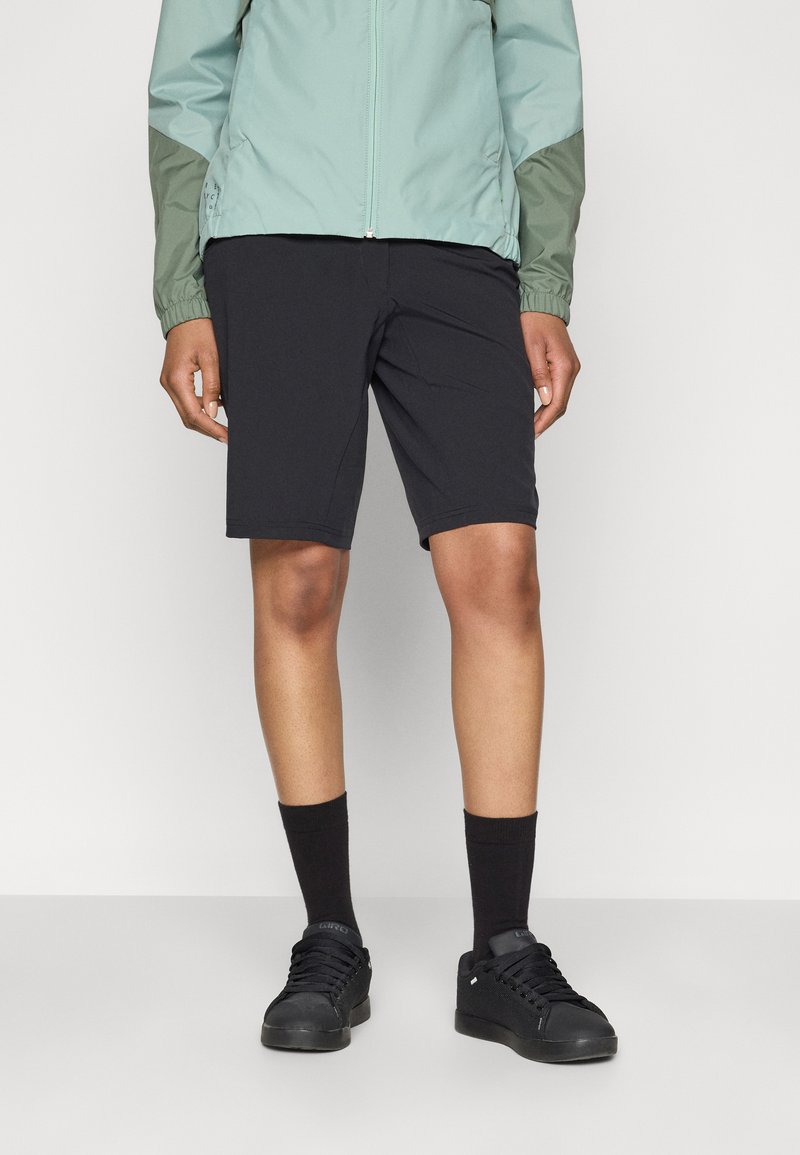 Shorts de sport noirs, en tissu léger, longueur au-dessus du genou. Portés avec des chaussettes noires et des baskets, ainsi qu'une veste vert menthe.