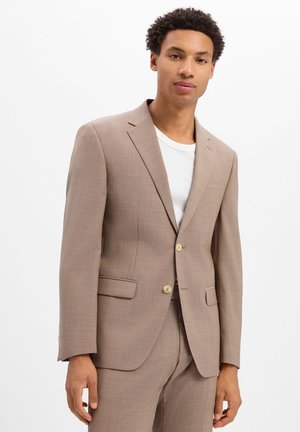 Junger Mann trägt eine beige Sakko und Hose über einem weißen T-Shirt mit Rundhalsausschnitt, steht vor einem schlichten weißen Hintergrund.