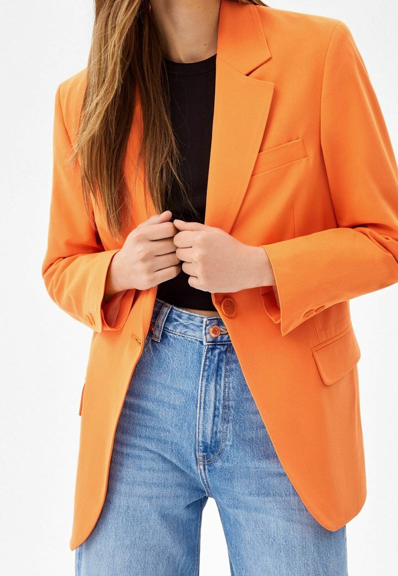 Oranžový strukturovaný blazer s ostrými klopami, zapínání na jeden knoflík a náprsní kapsou, v kombinaci s modrými džínami s vysokým pasem.