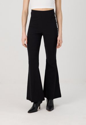 Mujer con pantalones acampanados negros de cintura alta y botines negros con tacón, combinados con una camiseta blanca corta, posando contra un fondo liso.