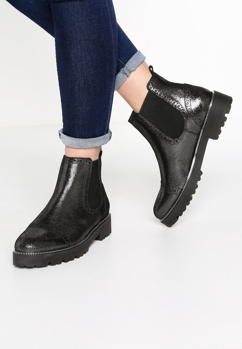 Botas de cuero negro brillante hasta el tobillo con suelas gruesas, llevadas con jeans de mezclilla oscura doblados, sobre un fondo blanco.
