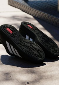 Baskets noires avec des rayures blanches montrées semelle vers le haut sur le trottoir, révélant des motifs sur la semelle et un détail de coussinet rouge près des talons.