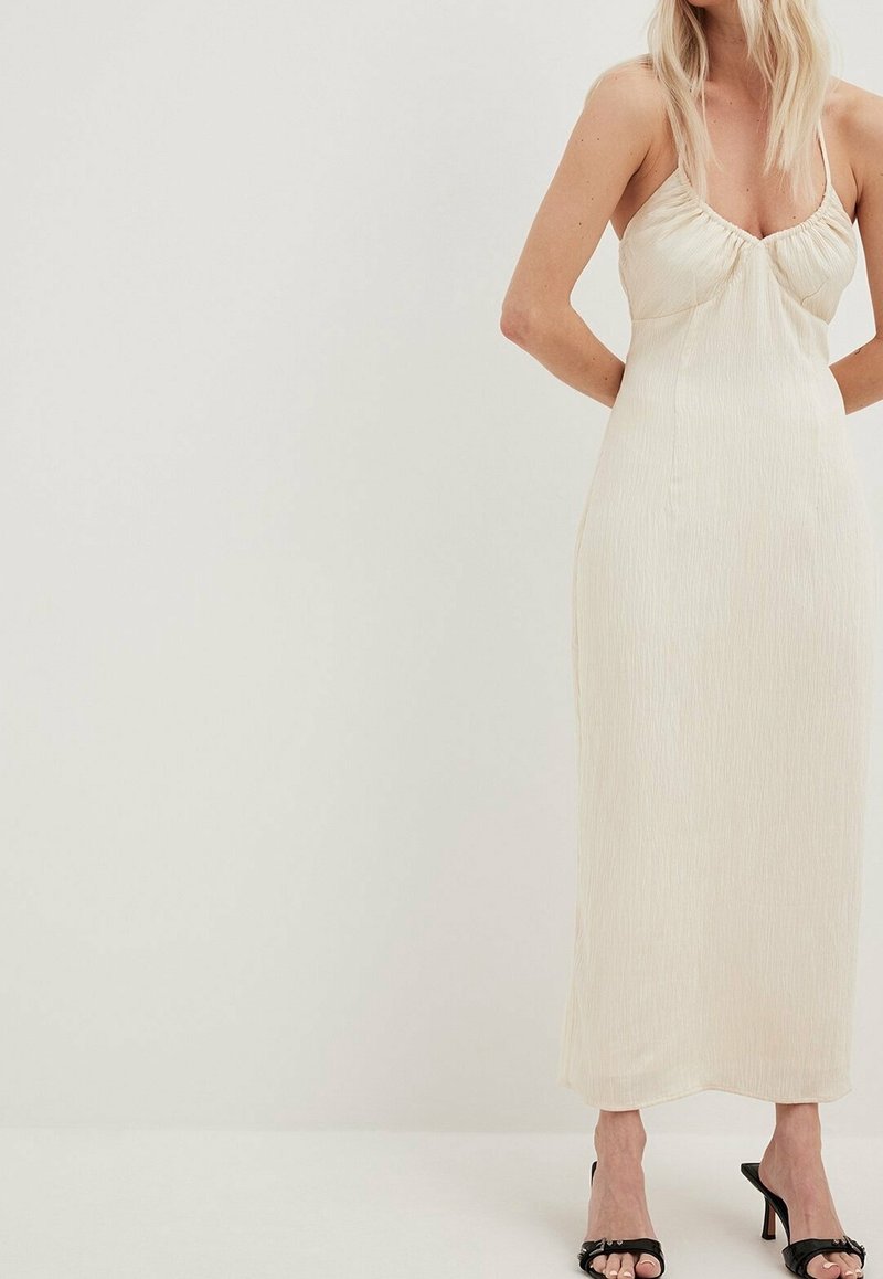 Femme portant une robe midi crème sans manches avec de fines bretelles et des talons ouverts noirs, debout sur un fond clair uni.